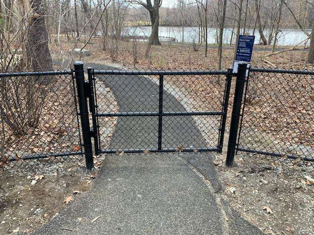 Chain link gate opening to a pathway near a water body, surrounded by bare trees and fallen leaves, illustrating residential chain link fencing options in Amesbury, MA.