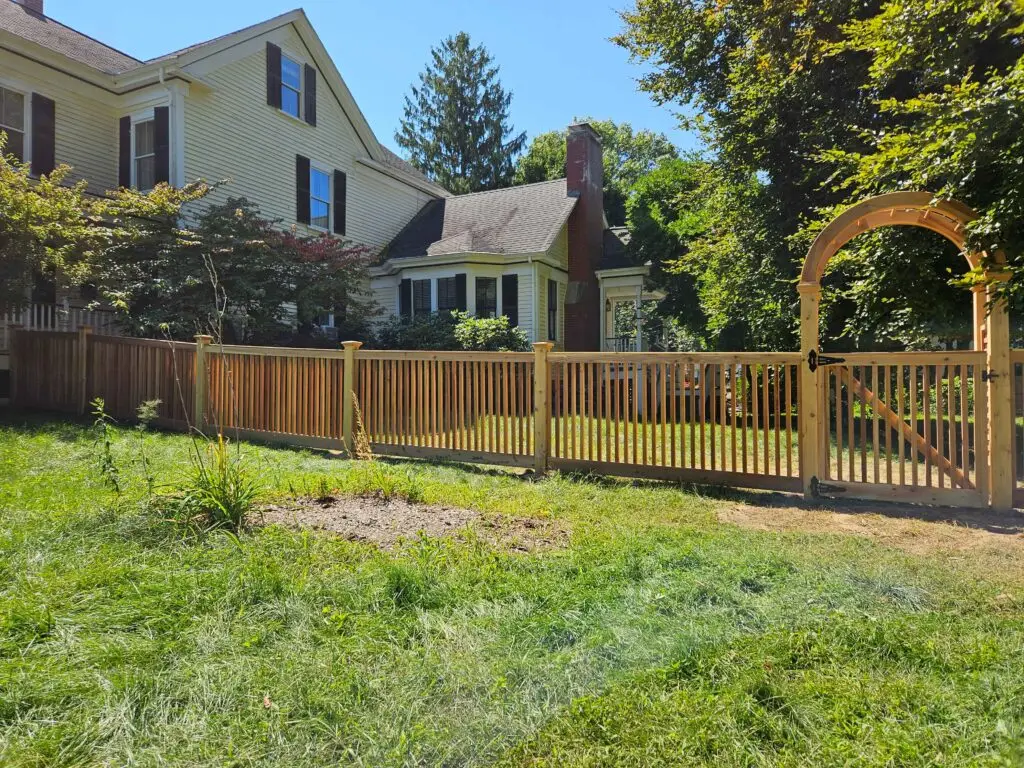 Custom wooden picket fence with arched gate, surrounded by lush greenery, enhancing residential curb appeal and showcasing Olympic Fence's quality craftsmanship.