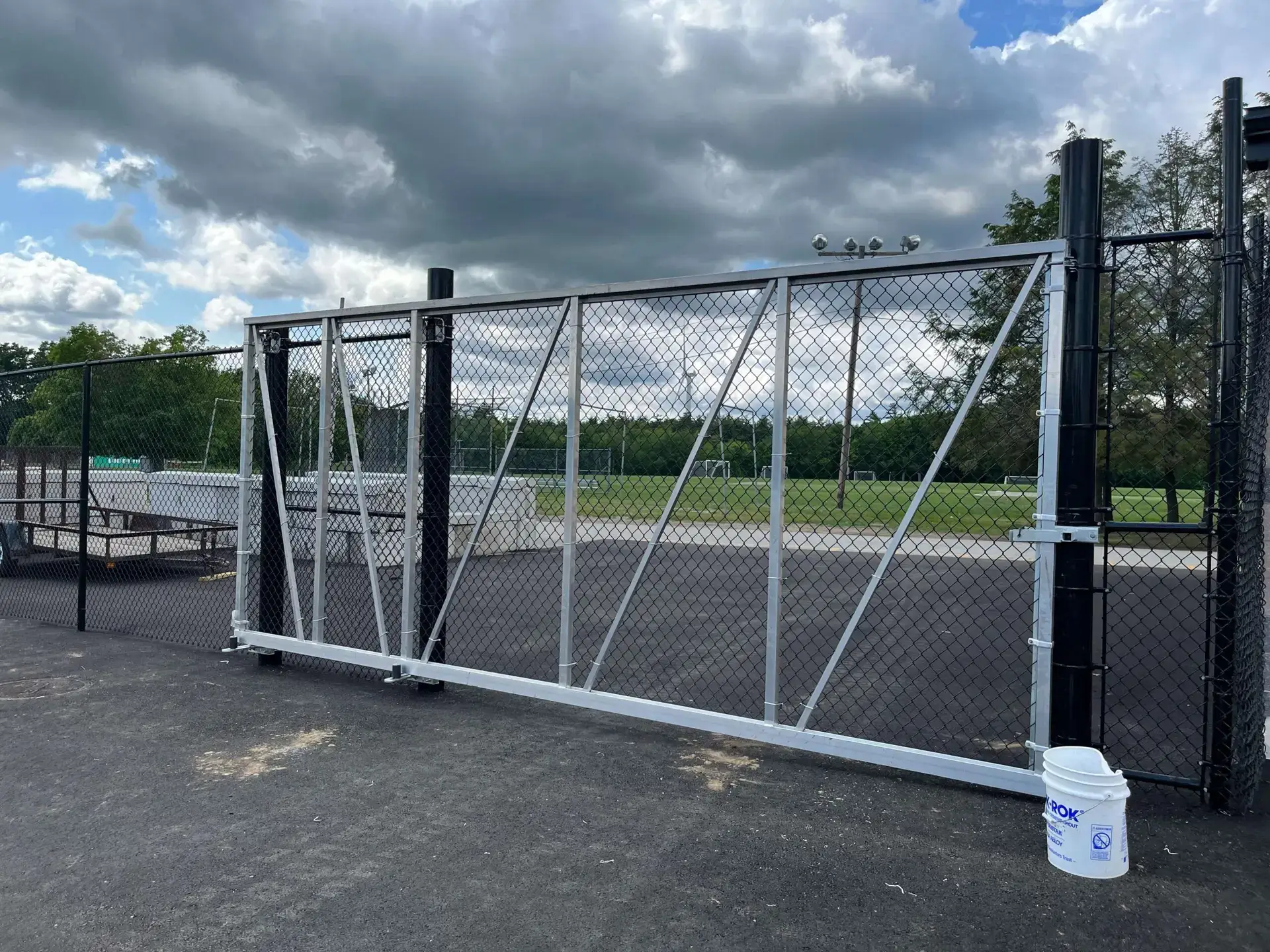 Sliding chain link gate with a black frame, leading to a grassy athletic field, surrounded by a chain link fence, showcasing durability for sports facilities.