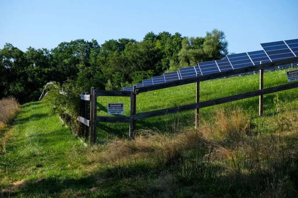 Solar panels on a fenced property with a "Danger: High Voltage" sign, surrounded by grass and trees, illustrating safety measures in fencing solutions.