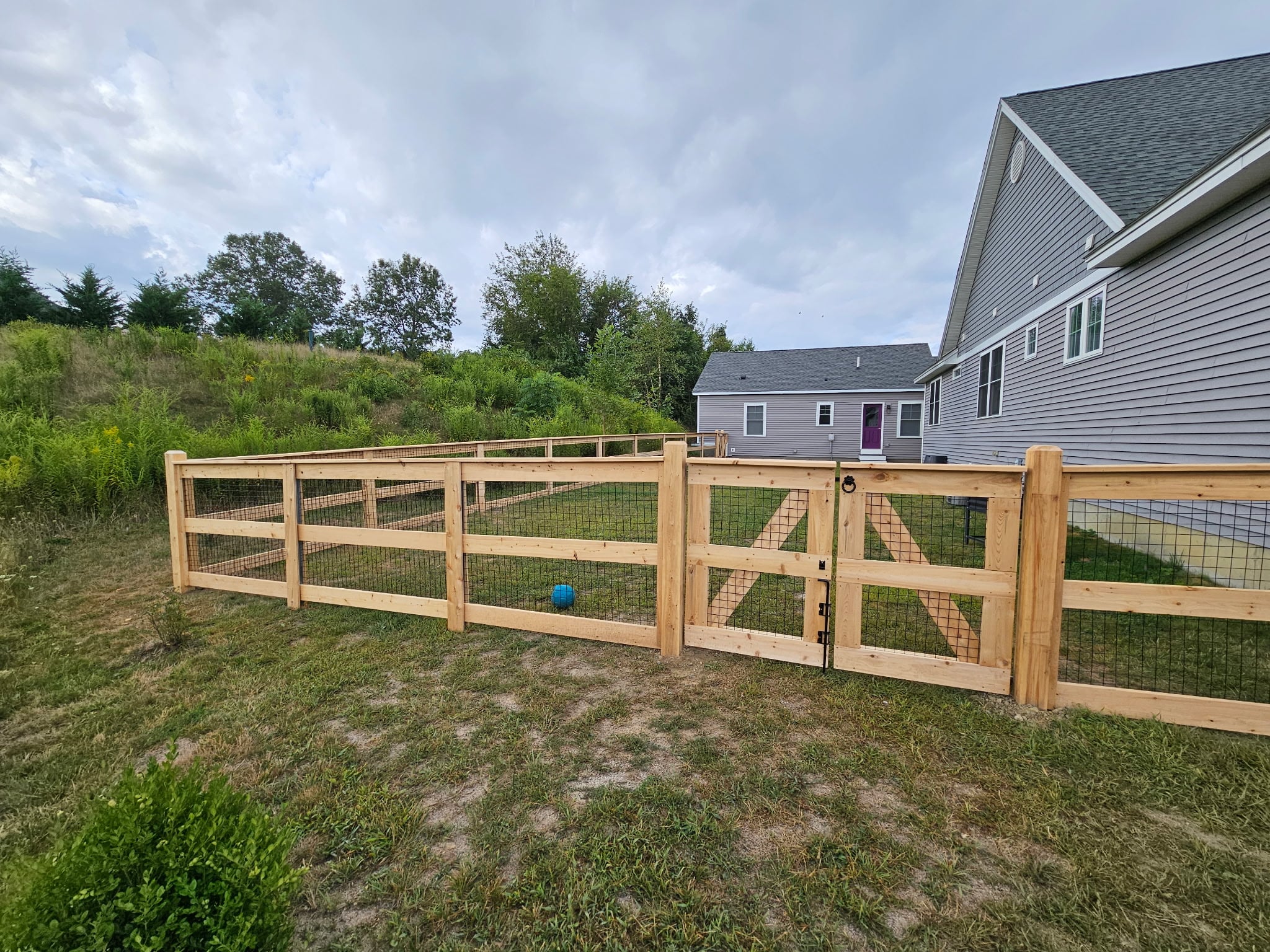 Cedar wood fenced yard with a gate, showcasing a budget-friendly fence installation in Amesbury, MA, surrounded by greenery and residential homes.
