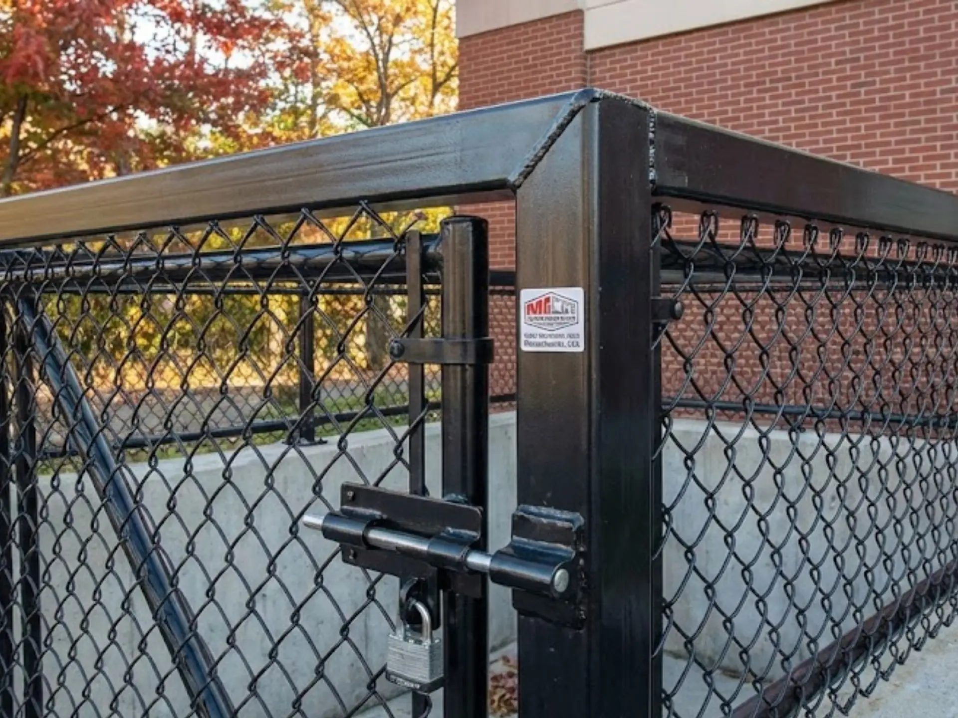 Chain link fence gate with lock, featuring heavy-duty construction and a black finish, set against a backdrop of autumn foliage and a brick building, illustrating security solutions for commercial and industrial properties in Amesbury.