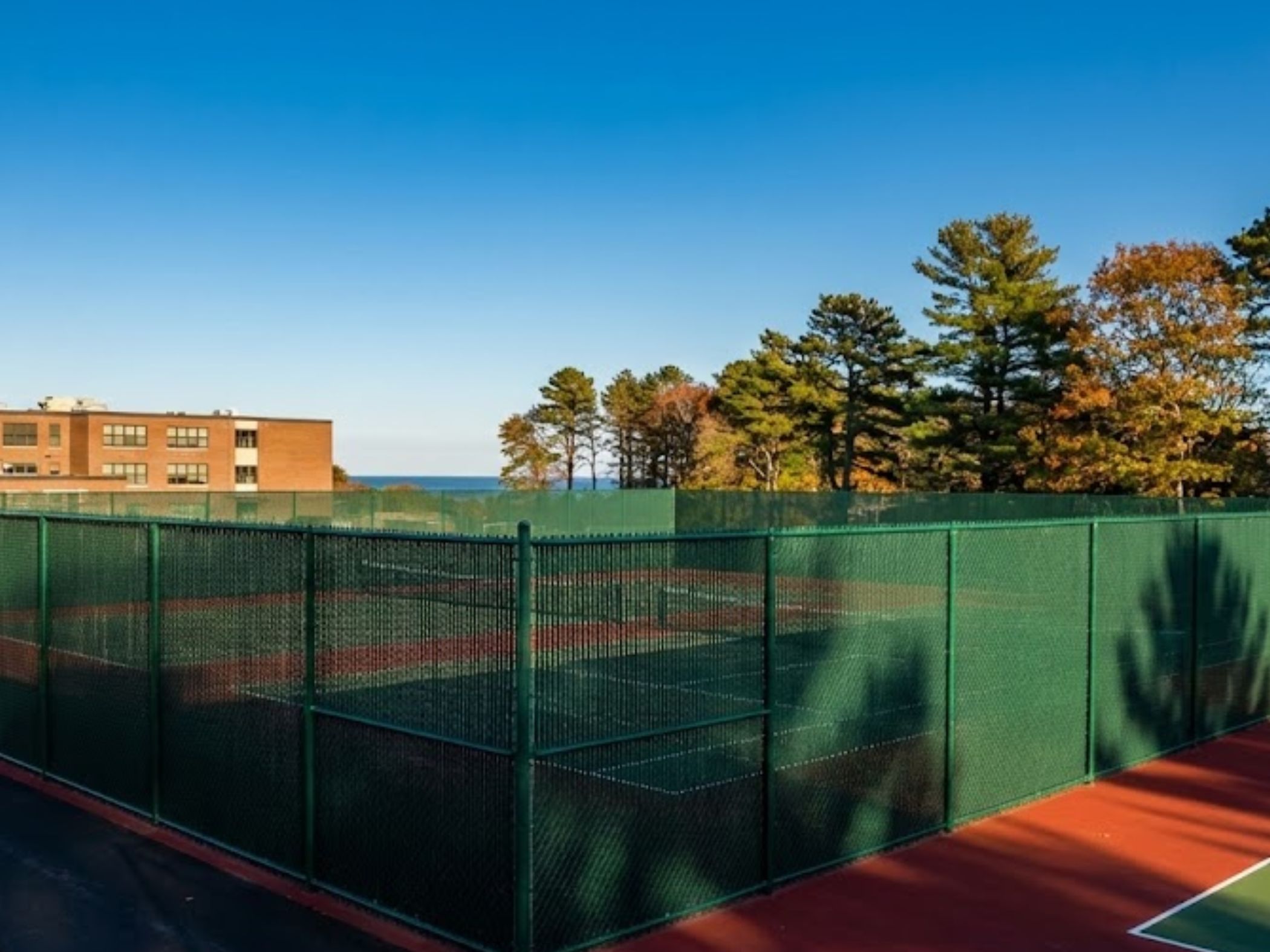 Tennis court enclosure featuring a 10-foot vinyl-coated chain-link fence, surrounded by trees and a school building, emphasizing commercial fencing solutions.