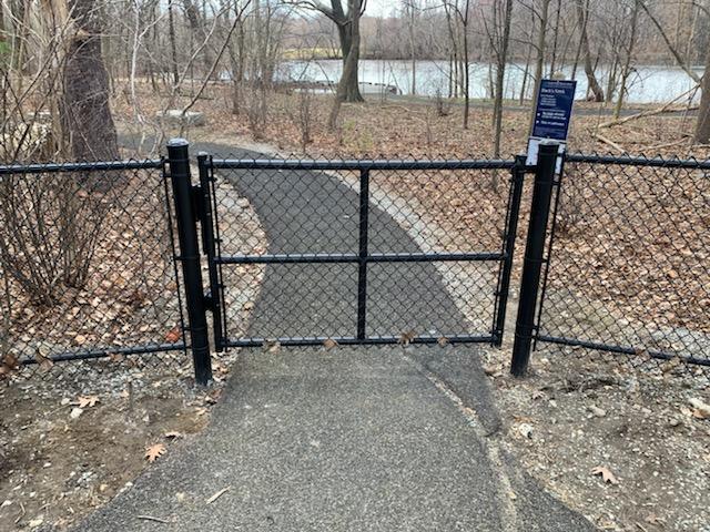 Black vinyl-coated chain link fence gate opening to a path in a wooded area near water, showcasing durable fencing options for properties in Newburyport.