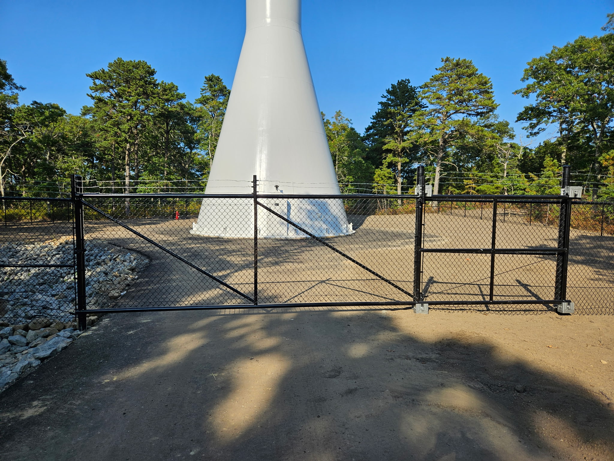 Black vinyl-coated chain link gate installed by Olympic Fence Co. in West Newbury, securing a gravel area near a white tower.