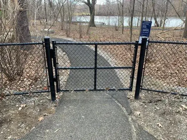 Chain link fence gate opening to a paved path in a wooded area, illustrating secure access for maintenance and safety.